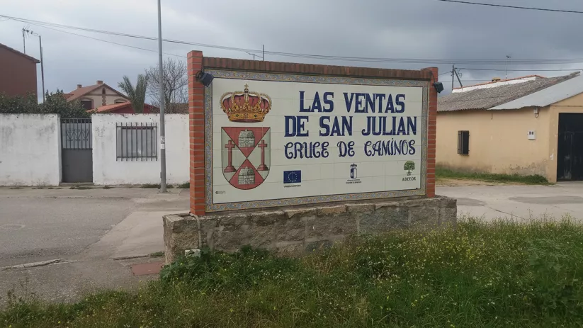 Mural de azulejos con un escudo de armas y letras grandes sobre un soporte de ladrillo y piedra, ubicado en una zona urbana bajo un cielo nublado.