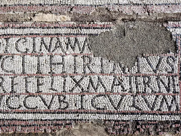 Detalle de mosaico antiguo con inscripción en teselas blancas y negras, parcialmente conservada.