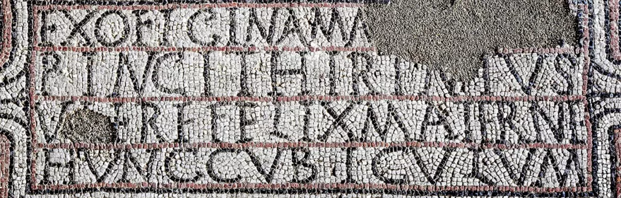 Detalle de mosaico antiguo con inscripción en teselas blancas y negras, parcialmente conservada.