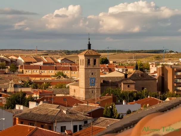 Panorámica aérea de los tejados de un pueblo con una torre de iglesia central, campos al fondo y nubes en el cielo.