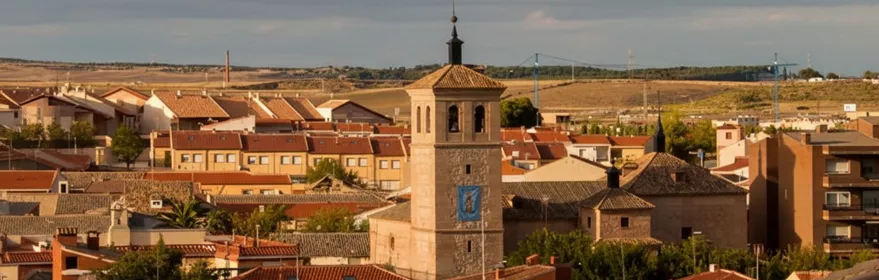 Panorámica aérea de los tejados de un pueblo con una torre de iglesia central, campos al fondo y nubes en el cielo.