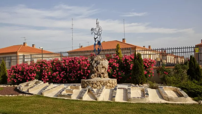 Una escultura metálica de una figura sobre un caballo encima de un pedestal de piedra, con flores rosas y un seto que forman la palabra "BIENVENIDO".