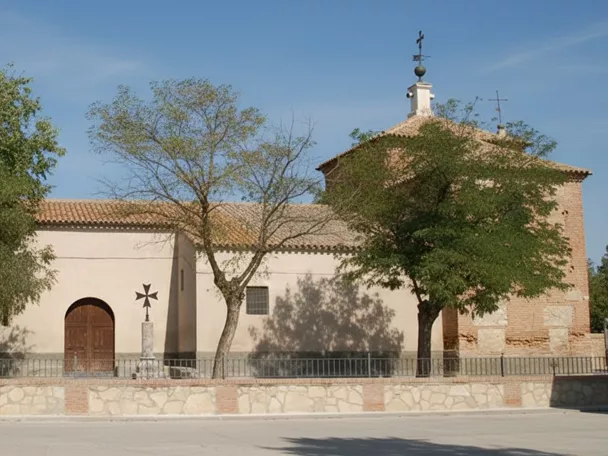 Vista frontal de una iglesia de una sola planta con paredes de color crema, una puerta de madera arqueada y un campanario de ladrillo con una cruz de hierro.