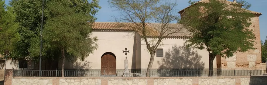 Vista frontal de una iglesia de una sola planta con paredes de color crema, una puerta de madera arqueada y un campanario de ladrillo con una cruz de hierro.