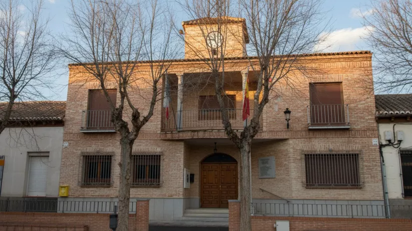 Fachada de ladrillo de un edificio de dos plantas con un reloj en una pequeña torre y banderas en un balcón central, flanqueada por árboles sin hojas.