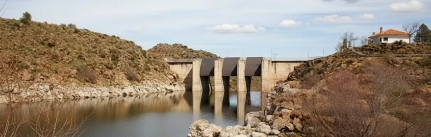 Una vista de un embalse con un fondo rocoso de colinas y una presa de concreto con cuatro compuertas, bajo un cielo azul con nubes dispersas, con un pequeño edificio en la colina a la derecha.