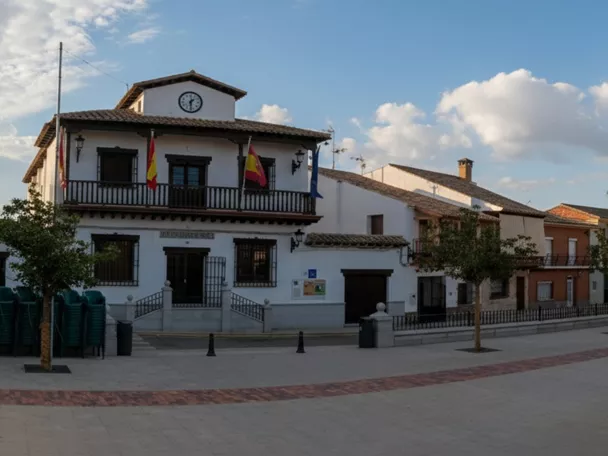 Una amplia vista panorámica de una plaza de pueblo vacía flanqueada por edificios históricos blancos y de ladrillo bajo un cielo parcialmente nublado.