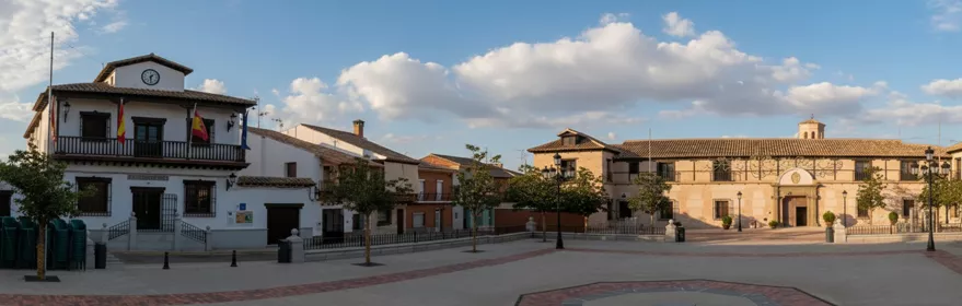 Una amplia vista panorámica de una plaza de pueblo vacía flanqueada por edificios históricos blancos y de ladrillo bajo un cielo parcialmente nublado.