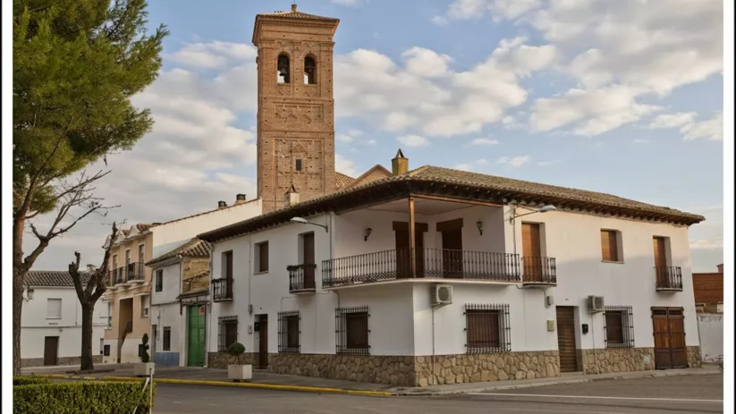 Una casa de pueblo blanca con balcones de hierro forjado y una torre de iglesia de ladrillo mudéjar que se eleva detrás, vista desde la esquina de una calle.