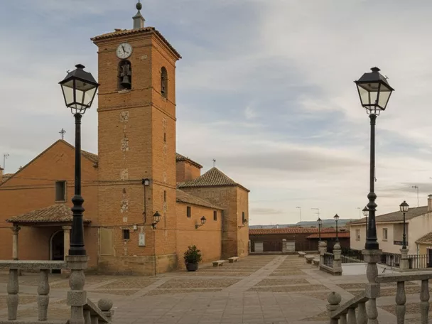 Una toma de ángulo bajo de una plaza adoquinada con farolas ornamentales, que muestra una iglesia de ladrillo con un campanario alto a la izquierda y un edificio municipal con banderas a la derecha.
