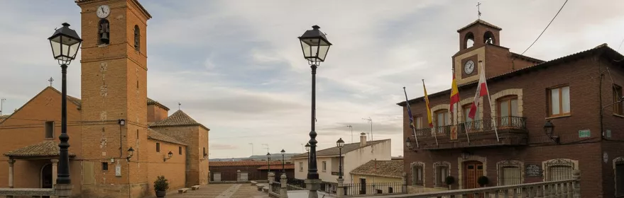 Una toma de ángulo bajo de una plaza adoquinada con farolas ornamentales, que muestra una iglesia de ladrillo con un campanario alto a la izquierda y un edificio municipal con banderas a la derecha.