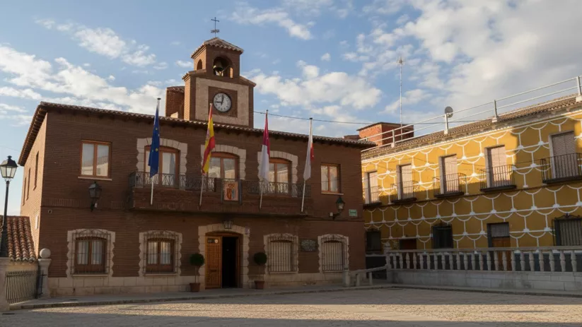 Un plano medio de la fachada de un edificio municipal de ladrillo rojo, que muestra un balcón con banderas, un reloj y un pequeño campanario sobre la entrada.
