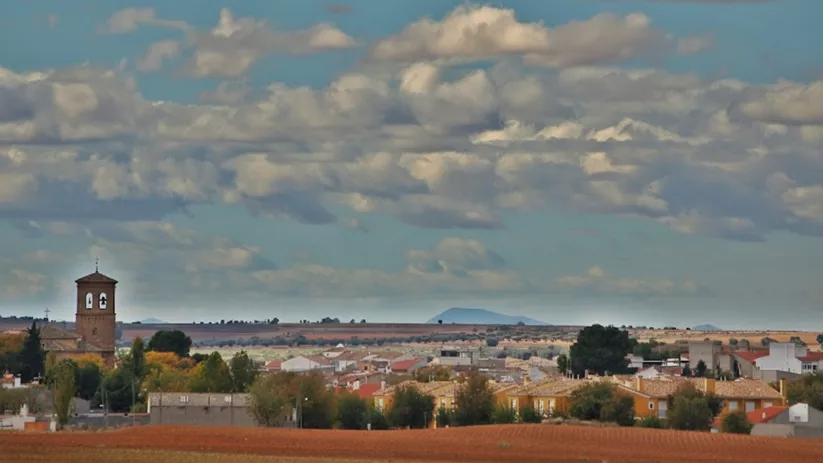 Un paisaje rural que muestra un pueblo con edificios bajos y un campanario de iglesia a la izquierda, bajo un cielo con muchas nubes.