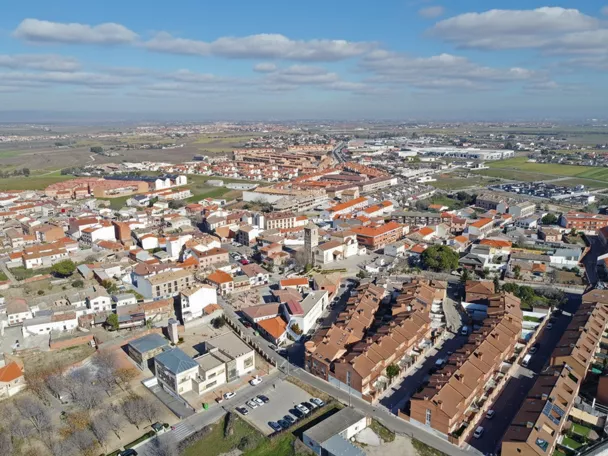 Una amplia vista panorámica de una plaza de pueblo vacía flanqueada por edificios históricos blancos y de ladrillo bajo un cielo parcialmente nublado.