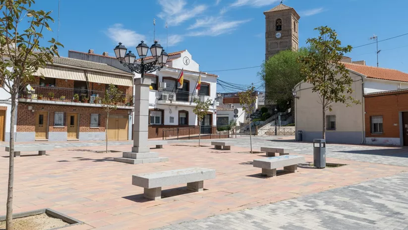 Plaza adoquinada con farolas ornamentales, que muestra una iglesia de ladrillo con un campanario alto a la izquierda y un edificio municipal con banderas a la derecha.