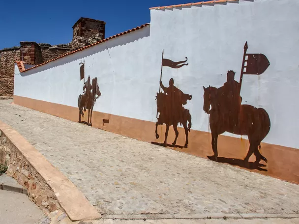 Siluetas metálicas de jinetes medievales en un muro blanco junto a una rampa empedrada.