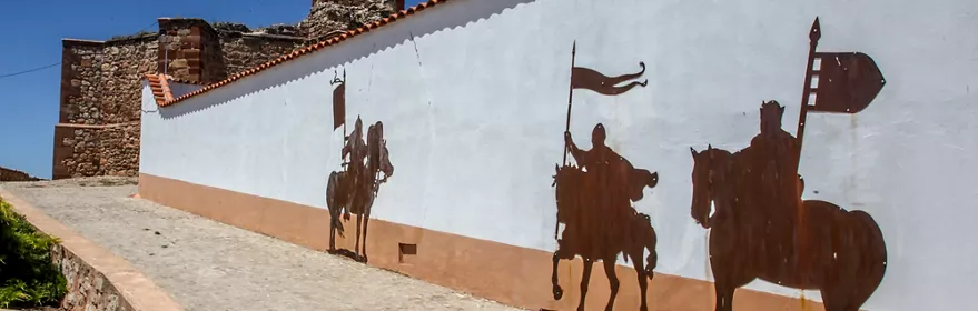 Siluetas metálicas de jinetes medievales en un muro blanco junto a una rampa empedrada.