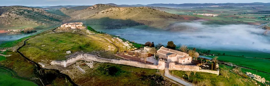 Vista aérea de fortaleza en colina rodeada de campos verdes y niebla baja