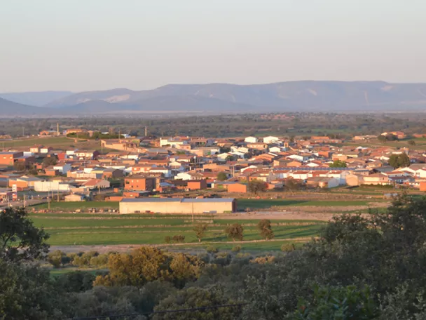 Vista panorámica de un pequeño pueblo rural con casas de tejados rojizos, rodeado de campos verdes y olivares, con colinas y montañas suaves al fondo bajo un cielo despejado.