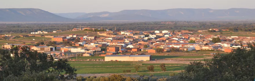 Vista panorámica de un pequeño pueblo rural con casas de tejados rojizos, rodeado de campos verdes y olivares, con colinas y montañas suaves al fondo bajo un cielo despejado.