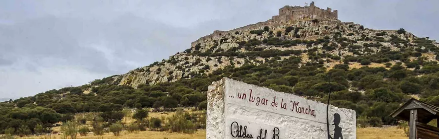 Paisaje rural con un castillo en lo alto de un cerro y un cartel que alude a “un lugar de La Mancha”.