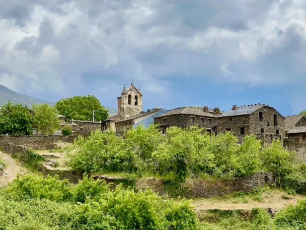 Conjunto de casas de piedra en ladera verde con torre y cielo nuboso.