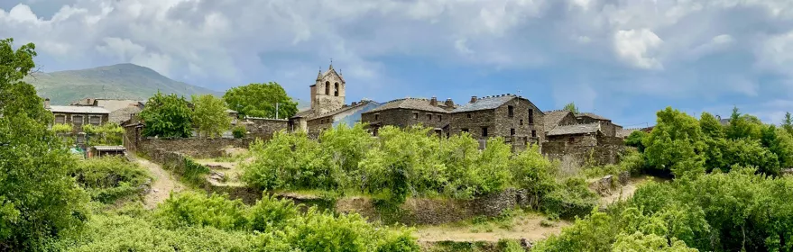 Conjunto de casas de piedra en ladera verde con torre y cielo nuboso.