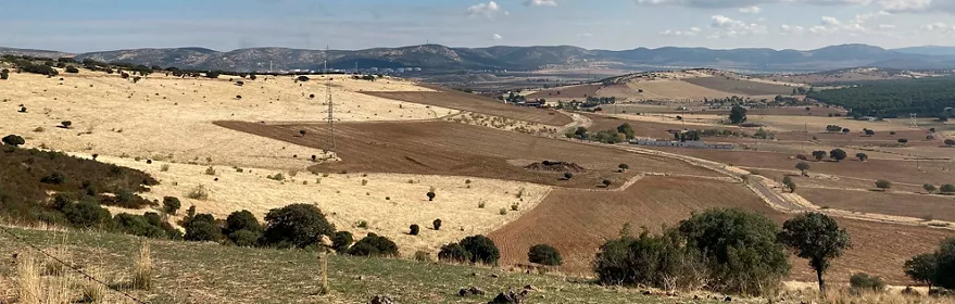 Paisaje de campos ondulados con tierras de cultivo y colinas al fondo bajo cielo azul con nubes.