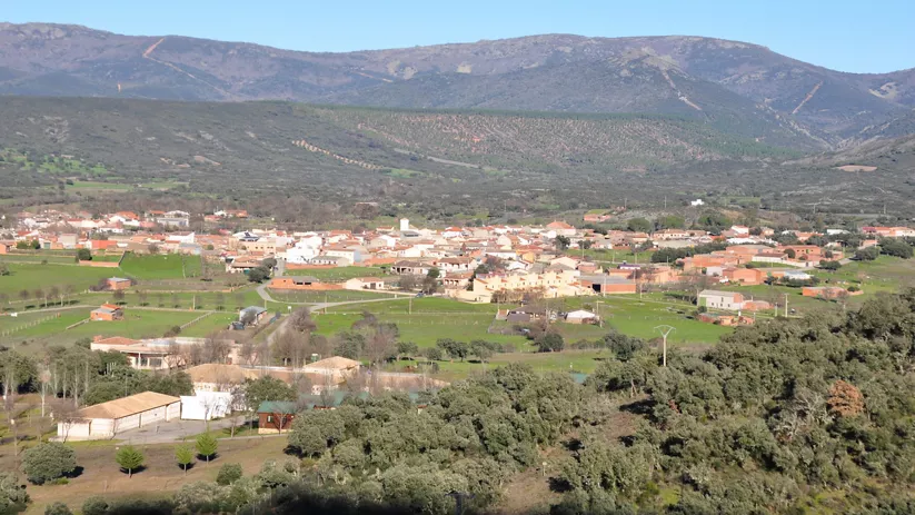 Vista aérea de un pueblo con campos verdes y montañas al fondo.