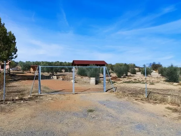 Acceso vallado a un recinto rural con edificaciones y terreno árido.
