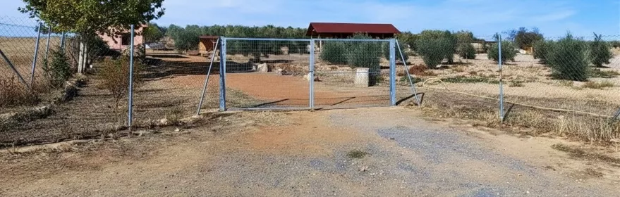Acceso vallado a un recinto rural con edificaciones y terreno árido.
