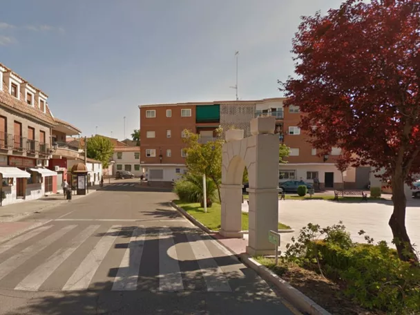 Vista de una calle urbana con un arco de piedra decorativo en primer plano, zonas verdes, árboles de hojas rojizas y edificios residenciales al fondo.