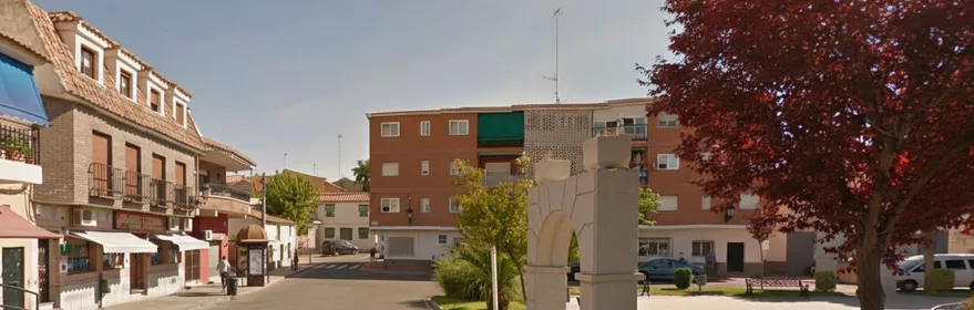 Vista de una calle urbana con un arco de piedra decorativo en primer plano, zonas verdes, árboles de hojas rojizas y edificios residenciales al fondo.