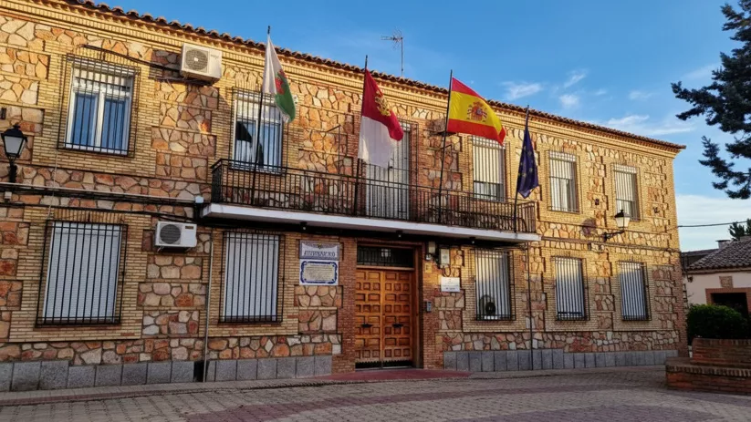 Edificio municipal de dos plantas con fachada de piedra rústica, balcones de forja y varias banderas oficiales ondeando sobre la entrada principal.