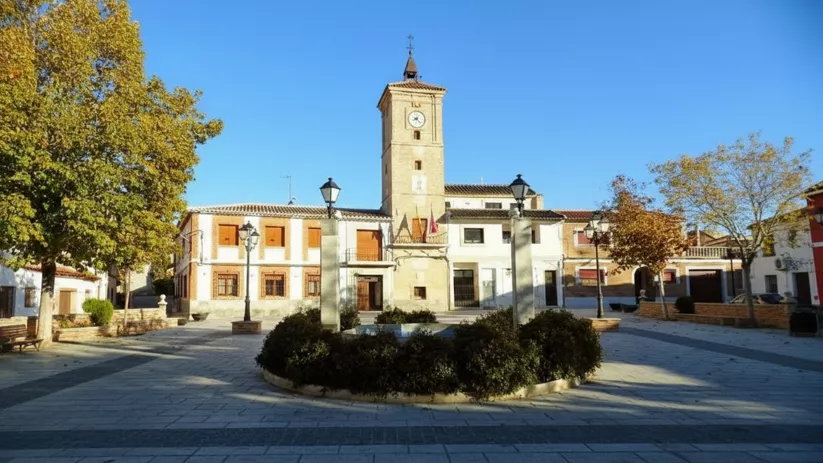 Una toma de nivel medio muestra una plaza de pueblo pavimentada con una torre de reloj central de ladrillo, un edificio de dos plantas y varios árboles con follaje otoñal.