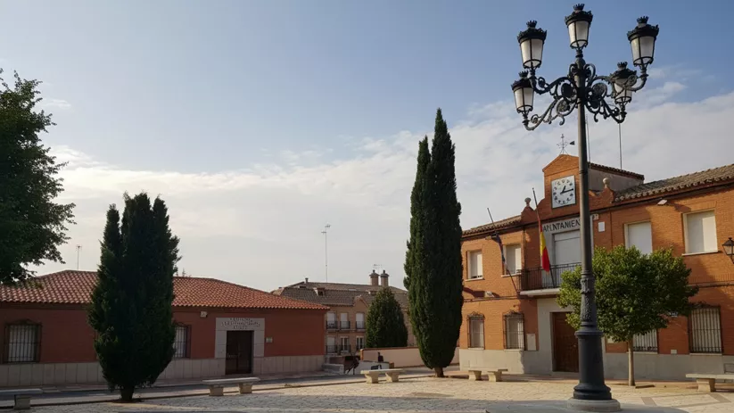 Vista de una plaza de pueblo pavimentada con piedra, con un gran farol de hierro forjado en el centro y edificios de ladrillo al fondo.