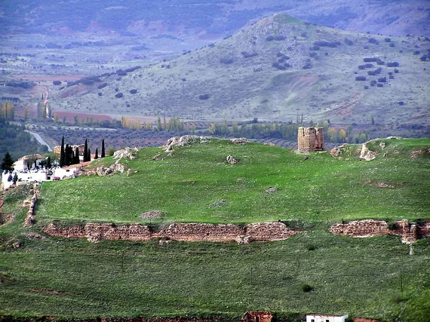 Vista panorámica de colina verde con restos de muralla y torre aislada.