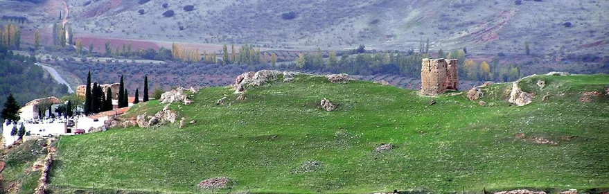 Vista panorámica de colina verde con restos de muralla y torre aislada.