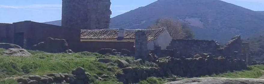 Torre de piedra en ruinas junto a edificaciones rurales y colinas al fondo.