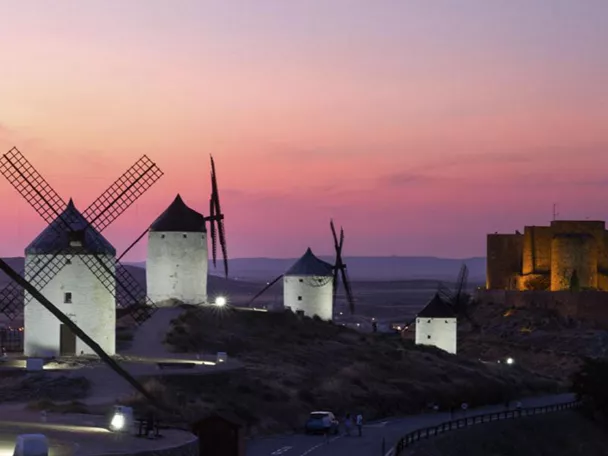 Molinos de viento iluminados al atardecer con fortaleza al fondo.