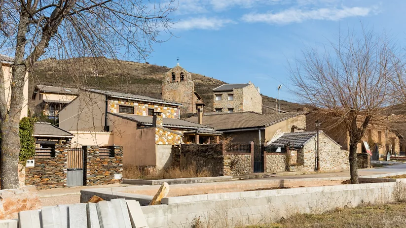 Calle de pueblo con casas rústicas, árboles sin hojas y torre al fondo