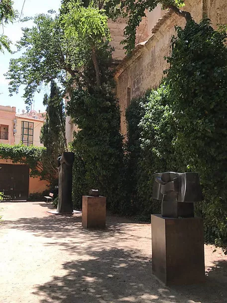 Sendero ajardinado con esculturas abstractas de metal entre muros cubiertos de hiedra.