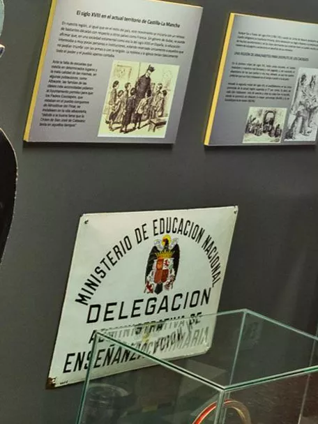 Sala expositiva con pupitres escolares antiguos y paneles informativos en las paredes.