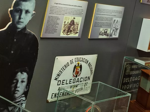 Sala expositiva con pupitres escolares antiguos y paneles informativos en las paredes.