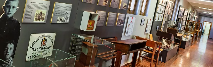Sala expositiva con pupitres escolares antiguos y paneles informativos en las paredes.