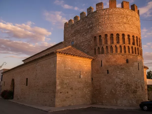Torre circular fortificada de piedra en Arenas de San Juan, con almenas y detalles de ladrillo, iluminada por la luz del atardecer.