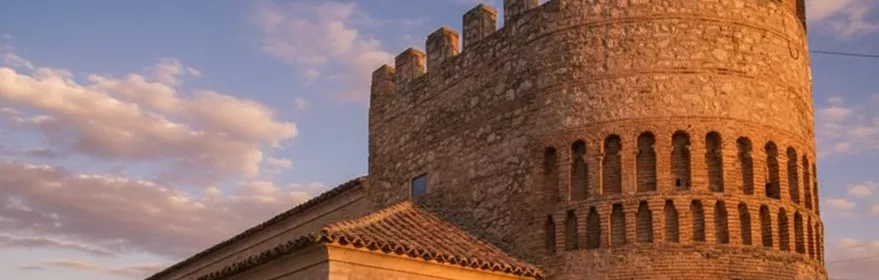 Torre circular fortificada de piedra en Arenas de San Juan, con almenas y detalles de ladrillo, iluminada por la luz del atardecer.