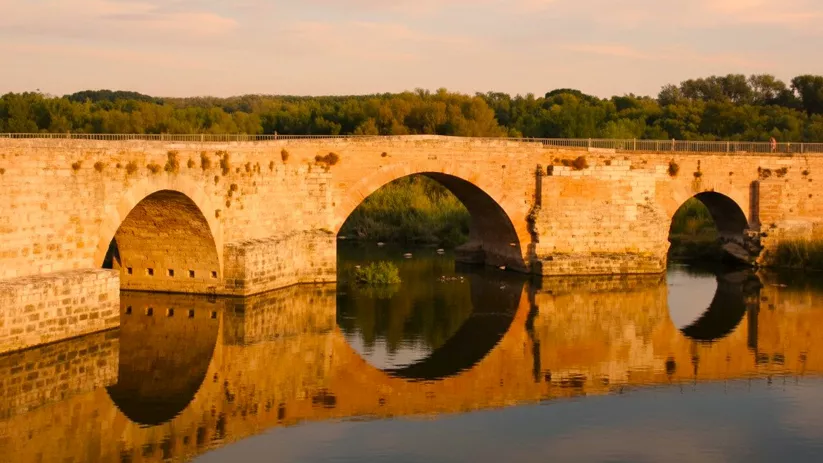 Puente de piedra iluminado por luz dorada, con reflejo circular en el agua.