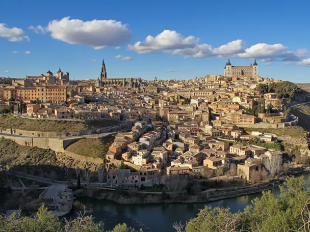 Una vista panorámica de una antigua ciudad de piedra situada en una colina sobre un río, bajo un cielo azul con nubes blancas.