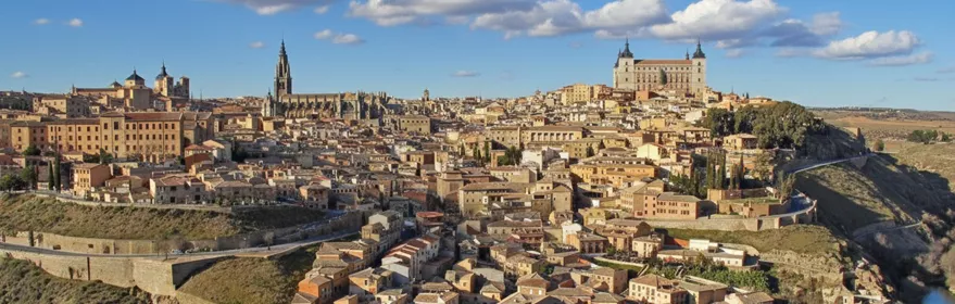 Una vista panorámica de una antigua ciudad de piedra situada en una colina sobre un río, bajo un cielo azul con nubes blancas.